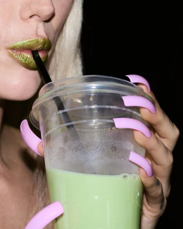 Close-up of Meredith Duxbury drinking a green beverage through a black straw, wearing green metallic lipstick and long pink acrylic nails.