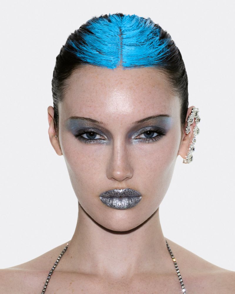 Close-up beauty portrait of model Meredith Duxbury with blue hair roots, metallic silver lips, and a crystal ear cuff against white.