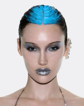 Close-up beauty portrait of model Meredith Duxbury with blue hair roots, metallic silver lips, and a crystal ear cuff against white.