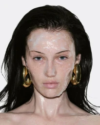 Close-up portrait of model Meredith Duxbury wearing glossy face cream and large gold earrings against a white background
