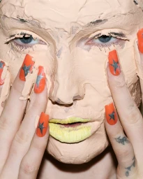 Close-up portrait of Meredith Duxbury with heavy beige foundation covering her face and bright yellow lips, framed by hands with orange star-patterned nails.