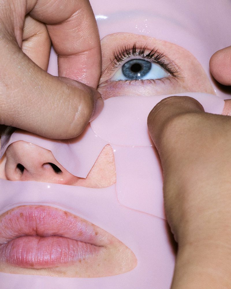 Close-up of hands applying a pink sheet face mask onto a model's face, focusing on her eye and lip area.