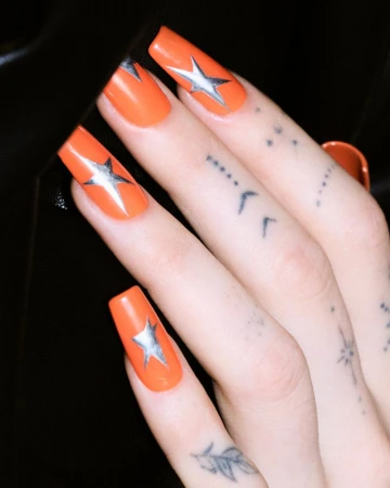 Close-up of long square orange fingernails featuring silver star nail art, with delicate tattoos visible on the fingers.