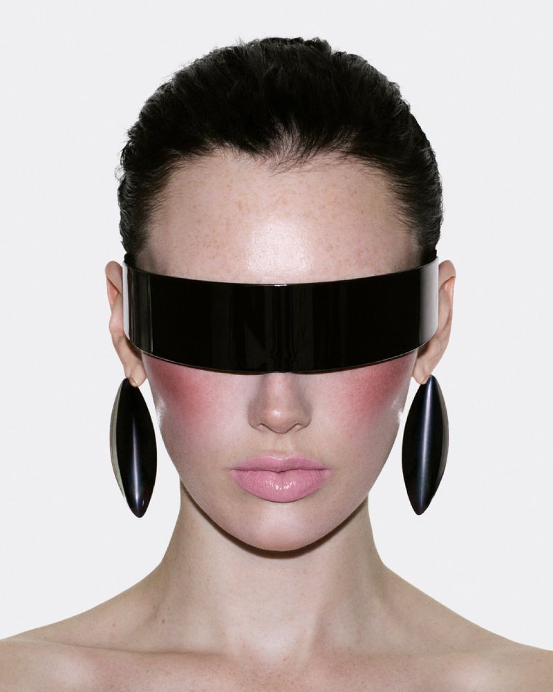 Close-up portrait of model Meredith Duxbury wearing black futuristic visor sunglasses and large teardrop earrings against a white background.