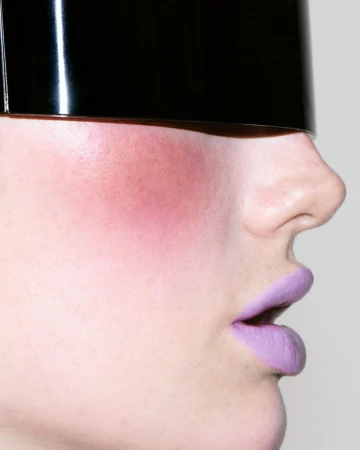 Close-up profile of Meredith Duxbury wearing a glossy black visor and vibrant purple lipstick against a plain white background