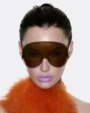 Close-up portrait of model Meredith Duxbury wearing oversized tinted visor sunglasses and an orange feathered collar.