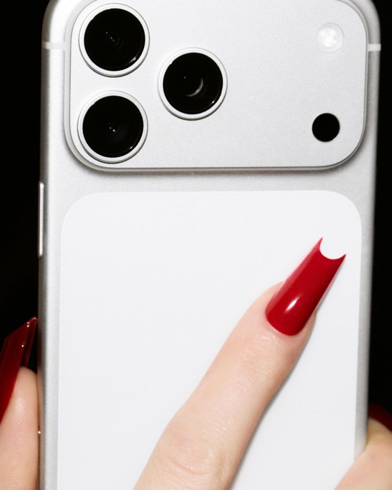 Close-up of a finger with glossy red long nails touching the back of a silver smartphone with three camera lenses against black.