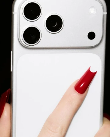 Close-up of a finger with glossy red long nails touching the back of a silver smartphone with three camera lenses against black.