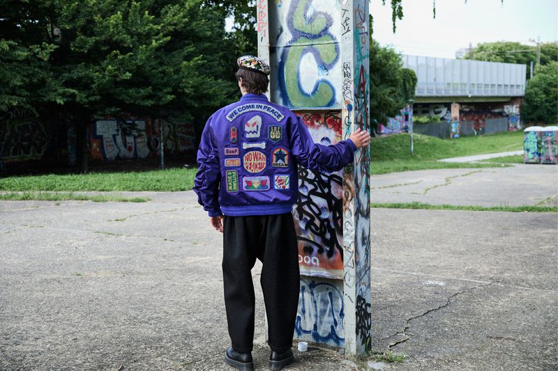 Person from the rear in a jacket with patches in a concrete outdoor setting with graffiti covered pillar, buildings, and trees.