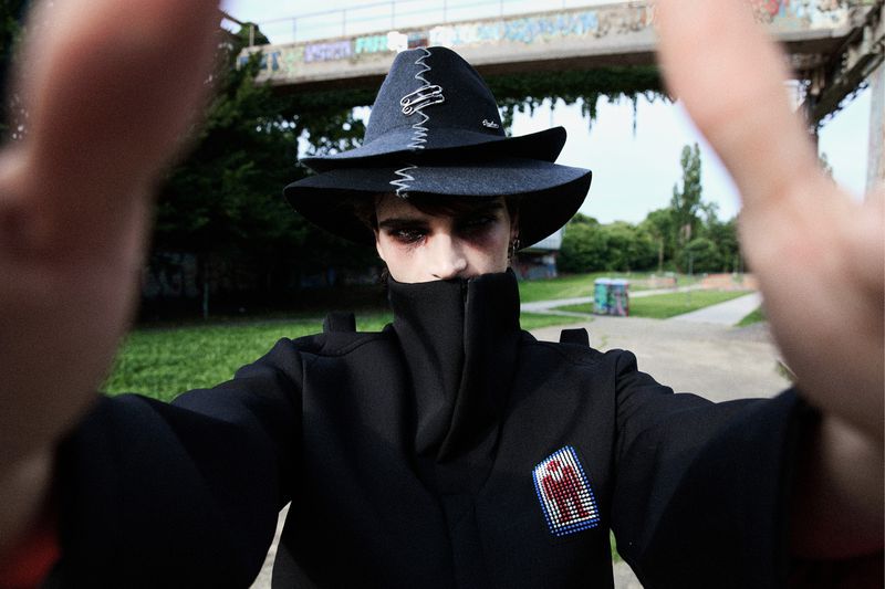 Close-up POV of a person in a black hat and clothing standing in a park with a bridge in the background, focusing on fashion and style.
