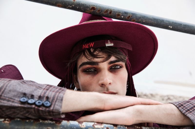 Close-up portrait of a young adult with heavy eye makeup, wearing a maroon hat and blazer, resting arms on a rusty metal bar.