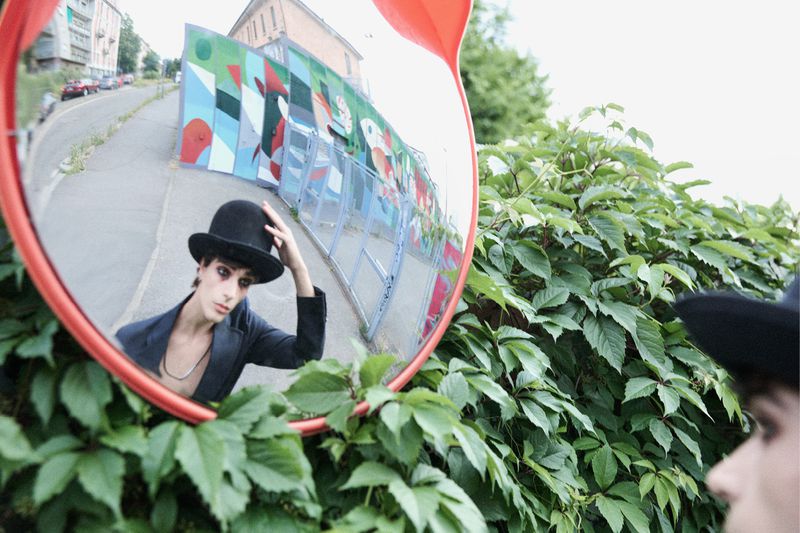 Reflection in a traffic mirror shows a person in a bowler hat and blazer standing among green plants in an urban outdoor setting.