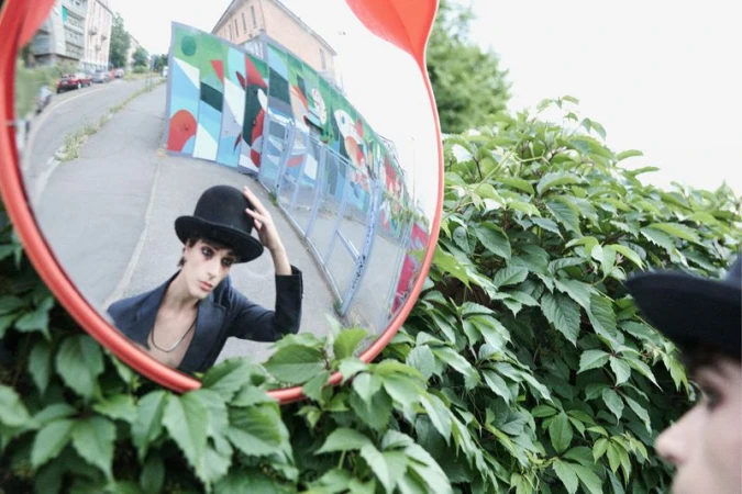 Reflection in a traffic mirror shows a person in a bowler hat and blazer standing among green plants in an urban outdoor setting.