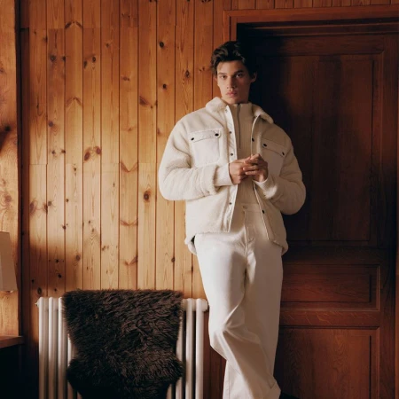 A male model wearing a white fleece jacket and trousers poses against a wood-paneled wall inside a chalet.