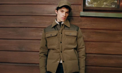 Model wearing an olive green quilted jacket and matching cap against horizontal wood paneling