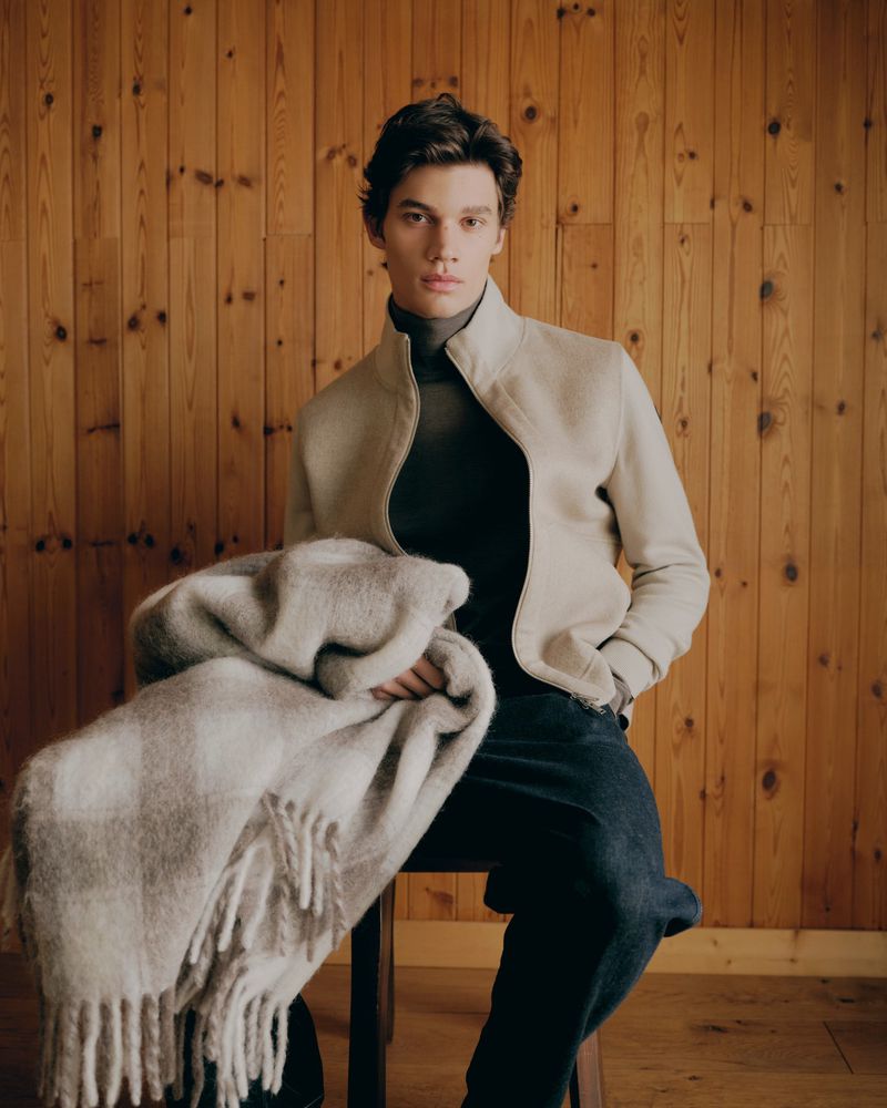 Male model holding a patterned wool blanket while seated against vertical wooden wall paneling