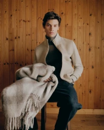 Male model holding a patterned wool blanket while seated against vertical wooden wall paneling