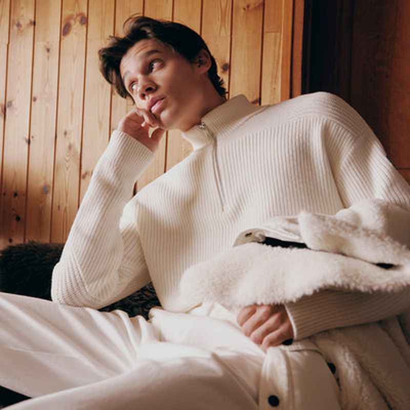 Male model in white zip-neck sweater resting chin on hand against a wooden wall interior