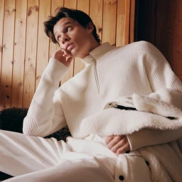 Male model in white zip-neck sweater resting chin on hand against a wooden wall interior