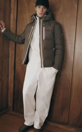 Male model in a dark puffer jacket, white trousers, and cap leaning against a wooden wall.
