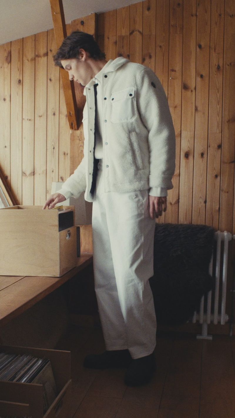 Model wearing a white fleece jacket and white pants standing in a wood-paneled interior