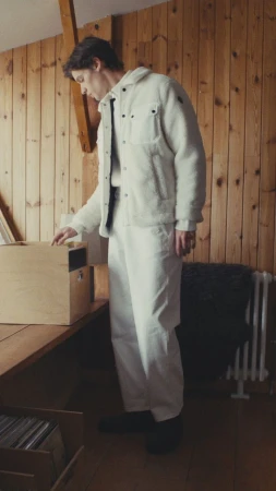 Model wearing a white fleece jacket and white pants standing in a wood-paneled interior