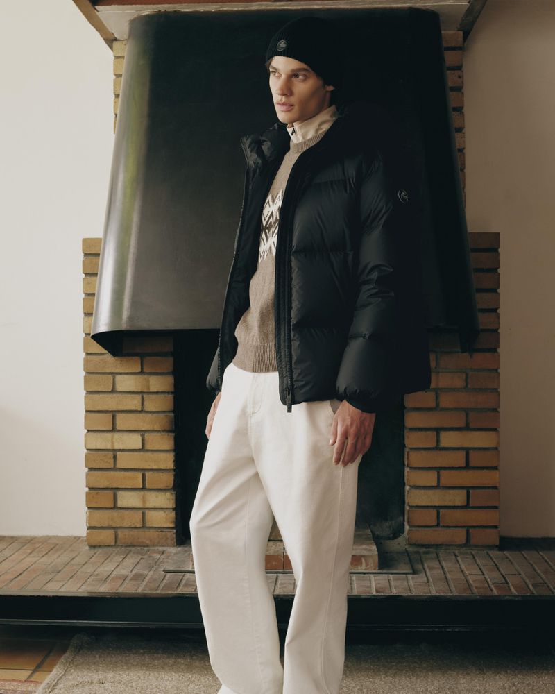 Model wearing a black puffer jacket and beanie standing indoors in front of a large brick fireplace.