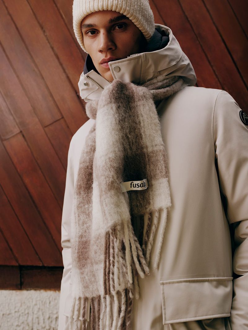 Close-up portrait of male model wearing a white winter jacket, matching beanie, and a chunky plaid scarf against wood paneling.
