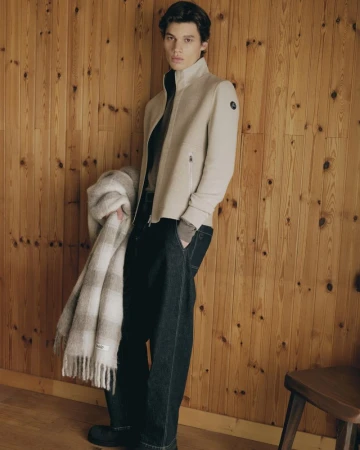 Male model in a cream zip-up jacket holding a plaid fringed blanket against wood paneling