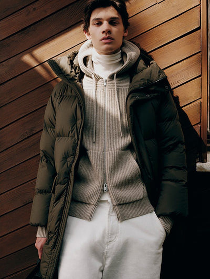 Male model wearing a quilted olive green puffer coat over a beige zip-up hoodie and white trousers against wooden panelling