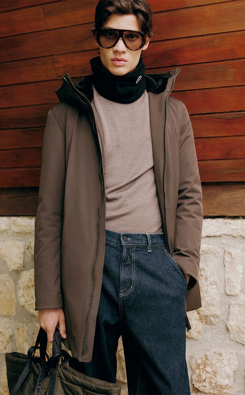 Frank De Beer wearing a brown winter coat, sunglasses, and dark trousers against wooden and stone walls.