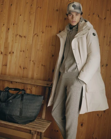 Model wearing white puffer jacket and grey tracksuit standing beside wooden bench with dark bag