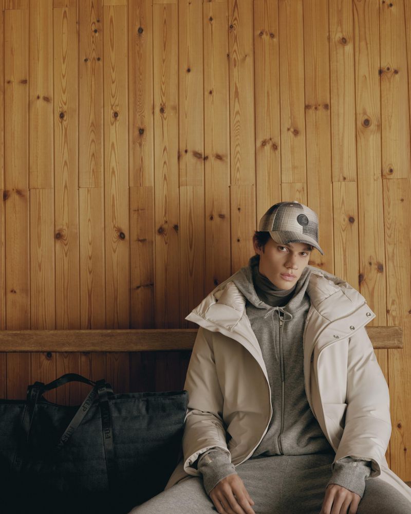 Male model seated against a vertical wooden panel wall wearing a cream puffer jacket, grey hoodie, and baseball cap