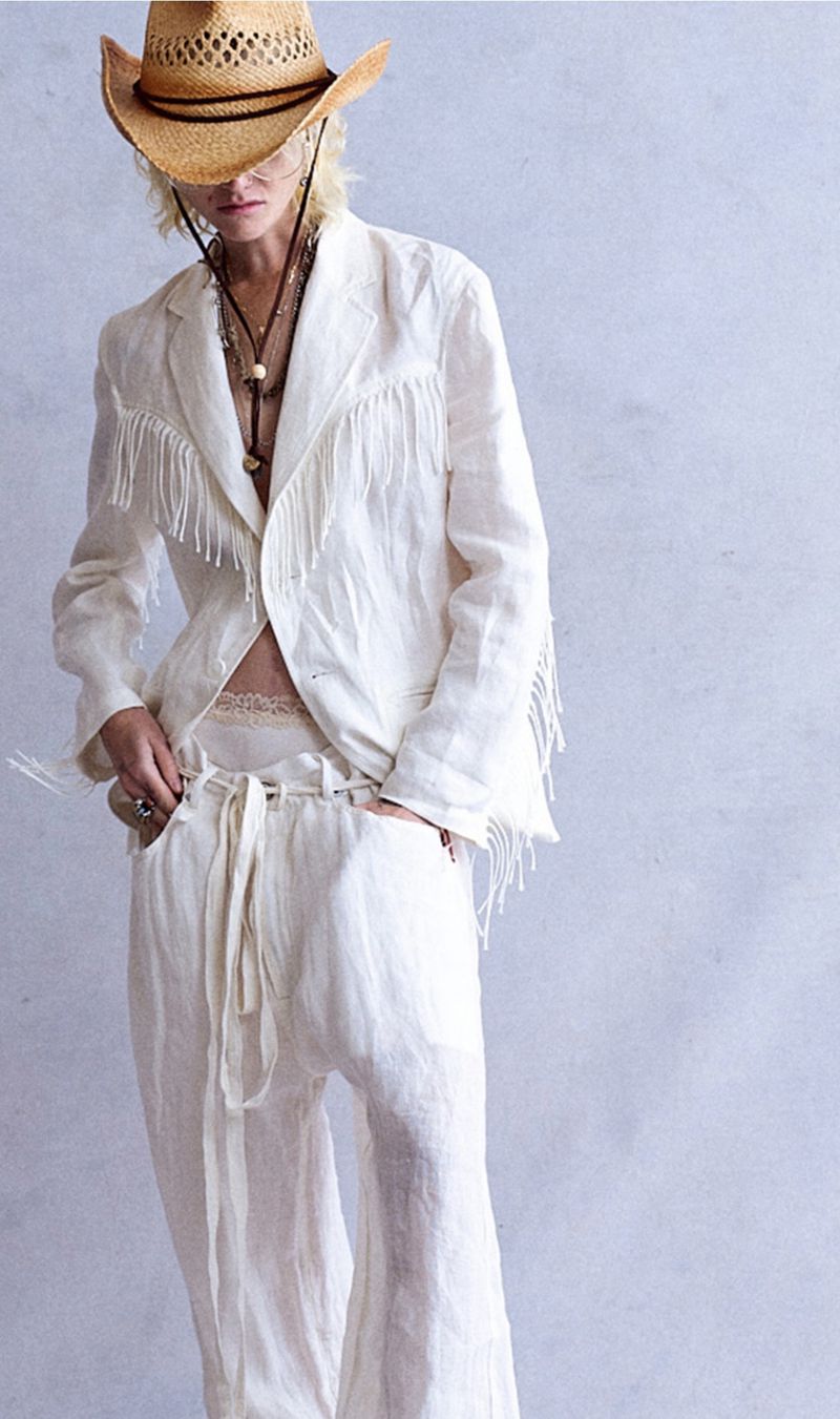Model wearing a white fringed jacket, matching trousers, and a straw cowboy hat against a textured wall.