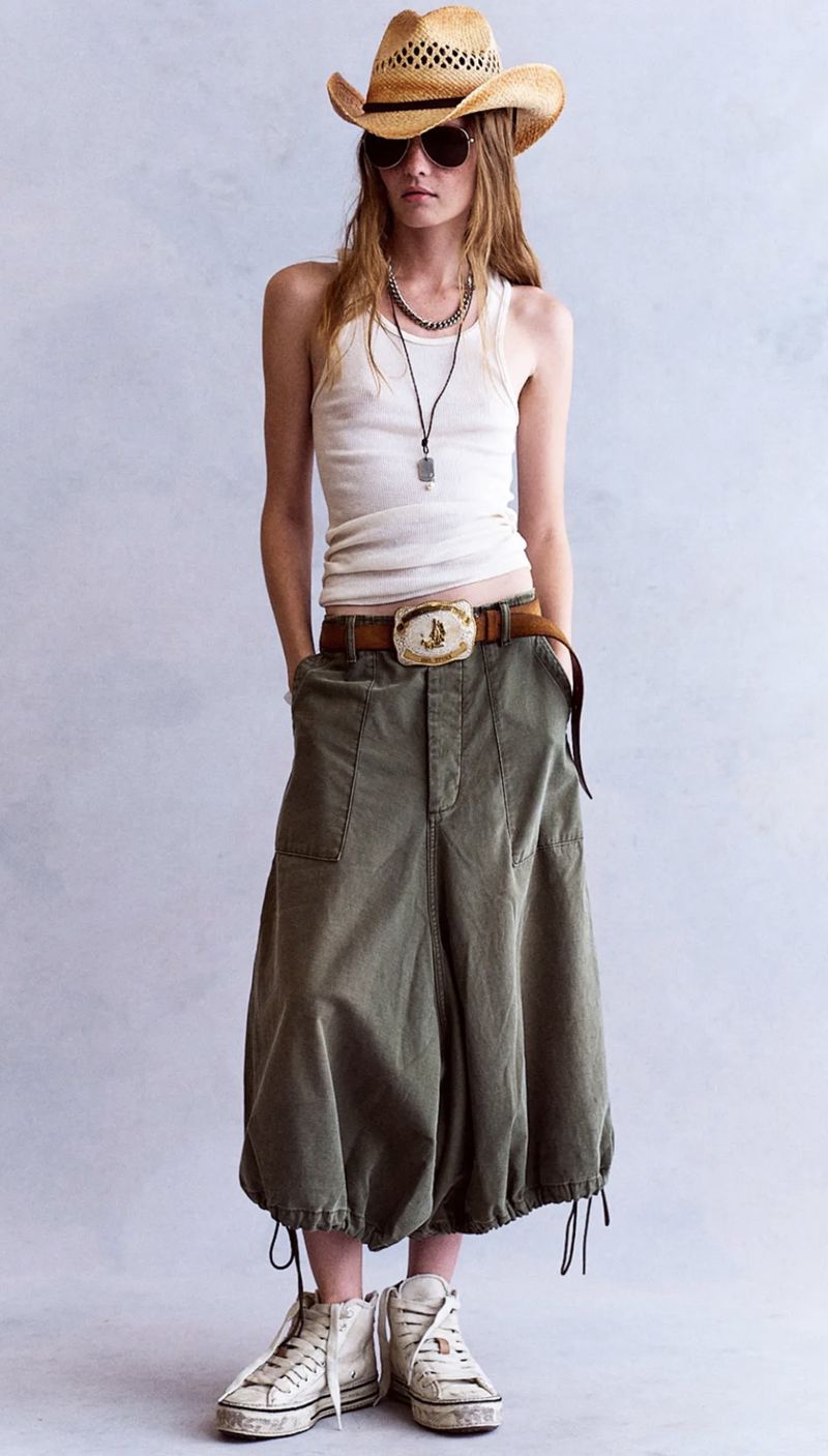 Model wearing a straw cowboy hat, white tank top, and olive utility pants against a plain grey backdrop