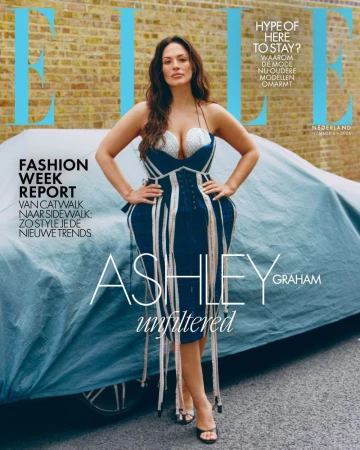 Model Ashley Graham standing in a structured blue dress leaning against a covered car in front of a brick wall