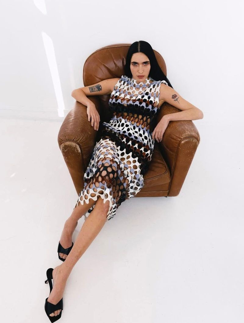 A fair-skinned young adult woman with long black hair poses in a brown leather armchair wearing a unique openwork dress and black mules.