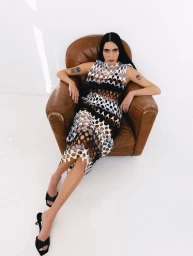 Model Valentine Valero seated in a brown leather armchair wearing a perforated patterned midi dress on a white studio background.