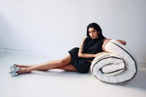 Model Valentine Valero in a black mini dress and silver heels reclining against a rolled white mattress on a white studio floor.