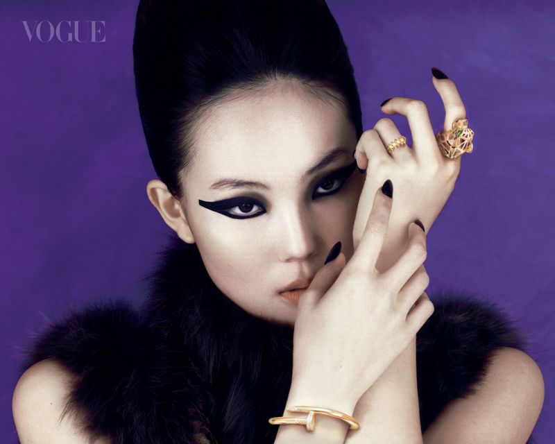 Model with dramatic winged eyeliner and statement jewelry posing against a purple background for Vogue Singapore