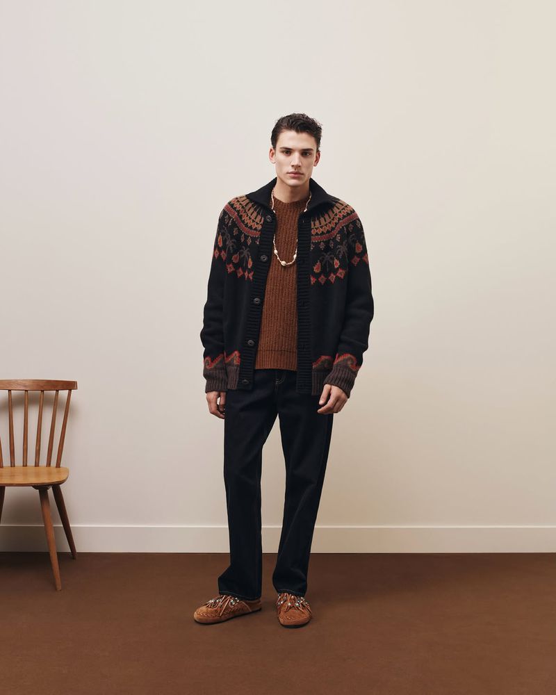 Model Tomasz Labis wearing a patterned cardigan and dark trousers standing in a studio with a wooden chair.