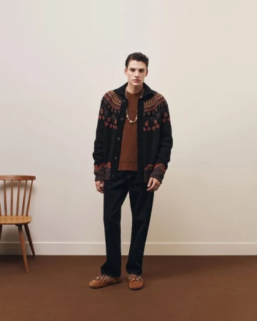Model Tomasz Labis wearing a patterned cardigan and dark trousers standing in a studio with a wooden chair.