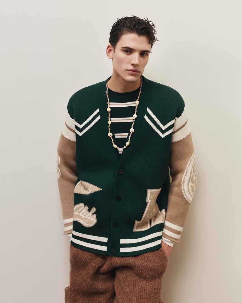 Model Tomasz Labis wearing a green patterned cardigan and brown trousers against a plain seamless background