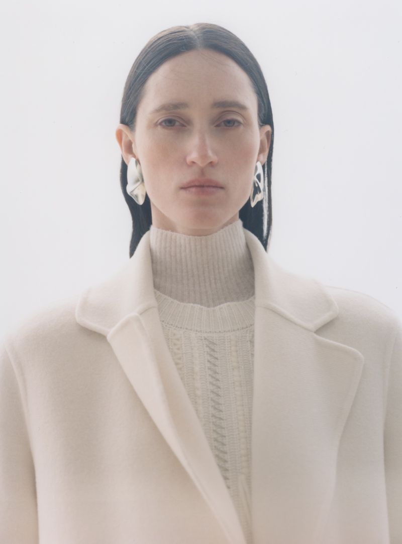 Model wearing a white coat and turtleneck with silver earrings against a plain background