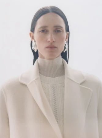 Model wearing a white coat and turtleneck with silver earrings against a plain background