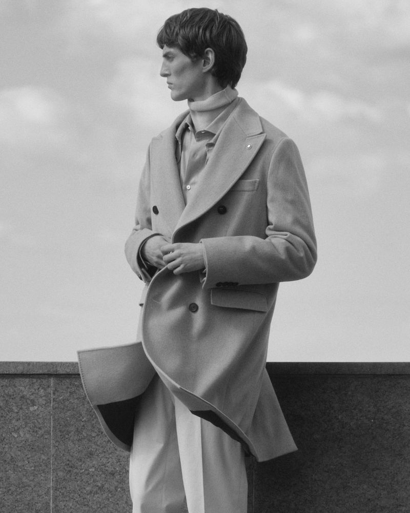 Monochrome portrait of a male model wearing a double-breasted coat and turtleneck against an open sky and low parapet.