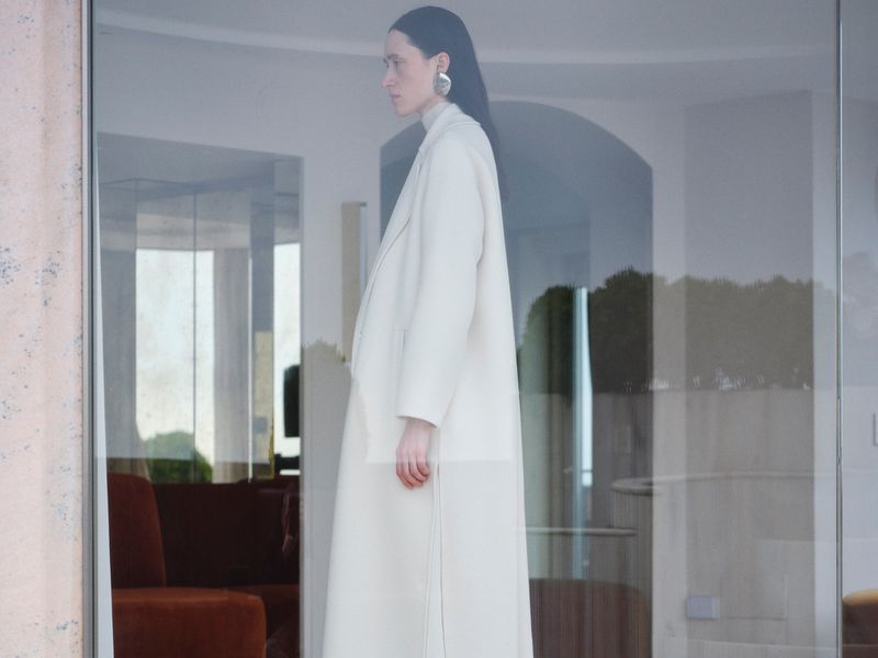 Model Helena Severin wearing a long cream coat viewed through a large glass window in a campaign for Peserico