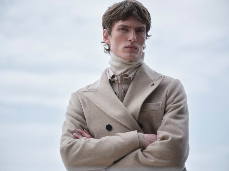 Male model with arms crossed wearing a cream wool coat and turtleneck against an overcast sky