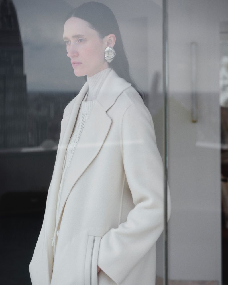 Model Helena Severin wearing a cream coat and turtleneck with silver earrings seen through a glass window.