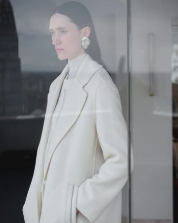 Model Helena Severin wearing a cream coat and turtleneck with silver earrings seen through a glass window.